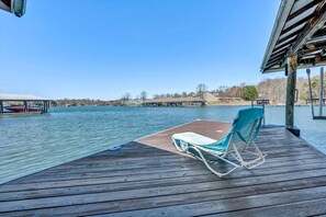 Eine Holzterrasse mit einem Liegestuhl und einem Tisch, mit Blick auf ruhiges Wasser, Boote und eine entfernte Küste.