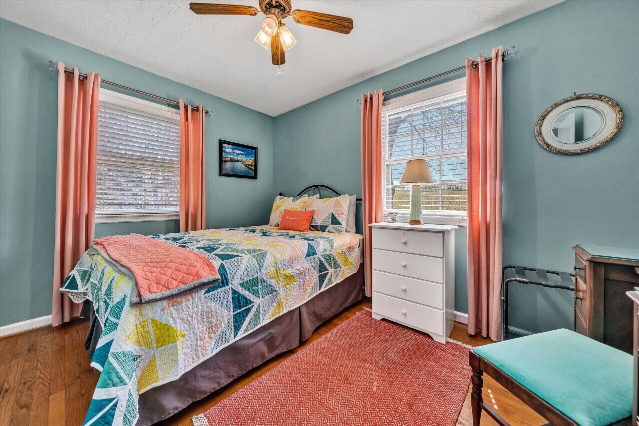 A bedroom with a bed, a dresser, a chair, a ceiling fan, and a window with curtains.
