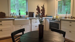 Una cocina moderna con armarios blancos, una mesa de comedor redonda y dos ventanas que dan a un bosque.