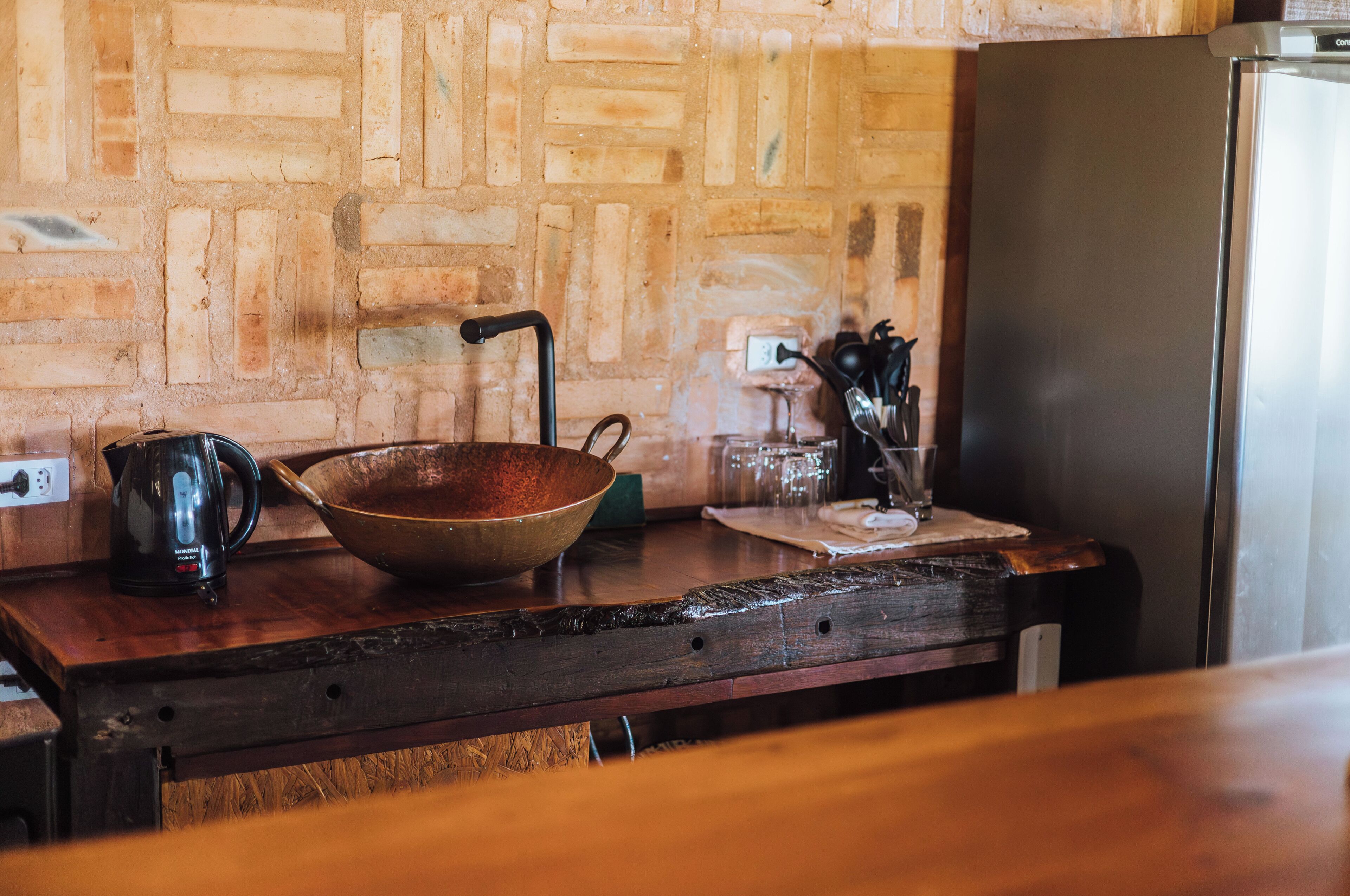 Une cuisine avec un mur en briques au style rustique, un plan de travail en bois, un réfrigérateur en acier inoxydable, une machine à café, un grand saladier et une bouilloire.