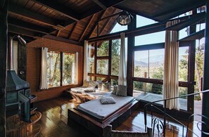 A rustic cabin with a wooden interior, a bed with pillows, a fireplace, and large windows offering a view of the outdoors. - Smart Hotel Reserva Santo Antônio do Pinhal (Santo Antonio do Pinhal)