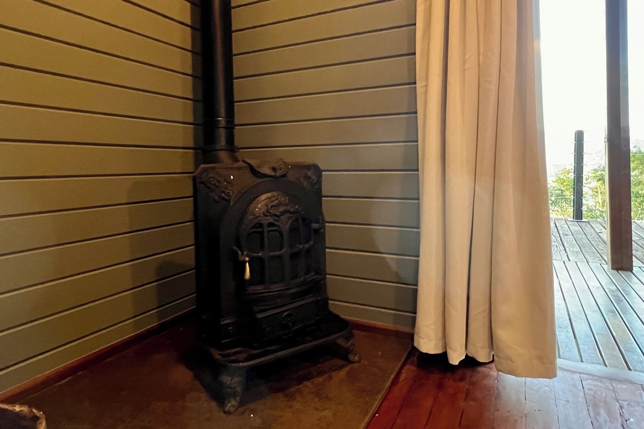 Una habitación de esquina con estufa de leña, piso de madera y ventana con cortinas.