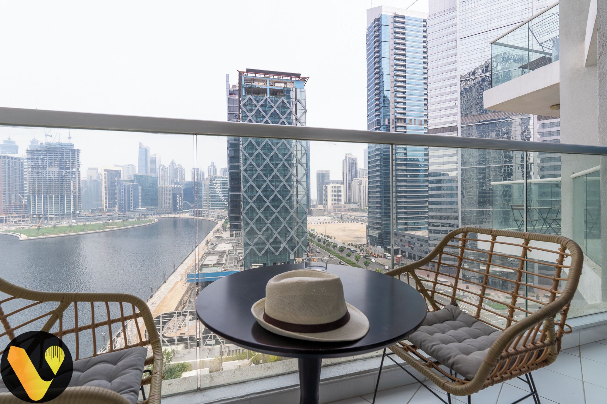 A balcony with wicker chairs, a table, and a hat. A cityscape with modern buildings and a river is visible in the background.
