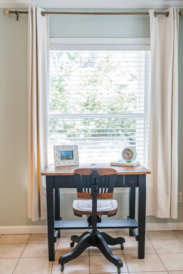 Un bureau avec une chaise devant une fenêtre munie de stores, un cadre photo et une horloge posée sur le bureau.