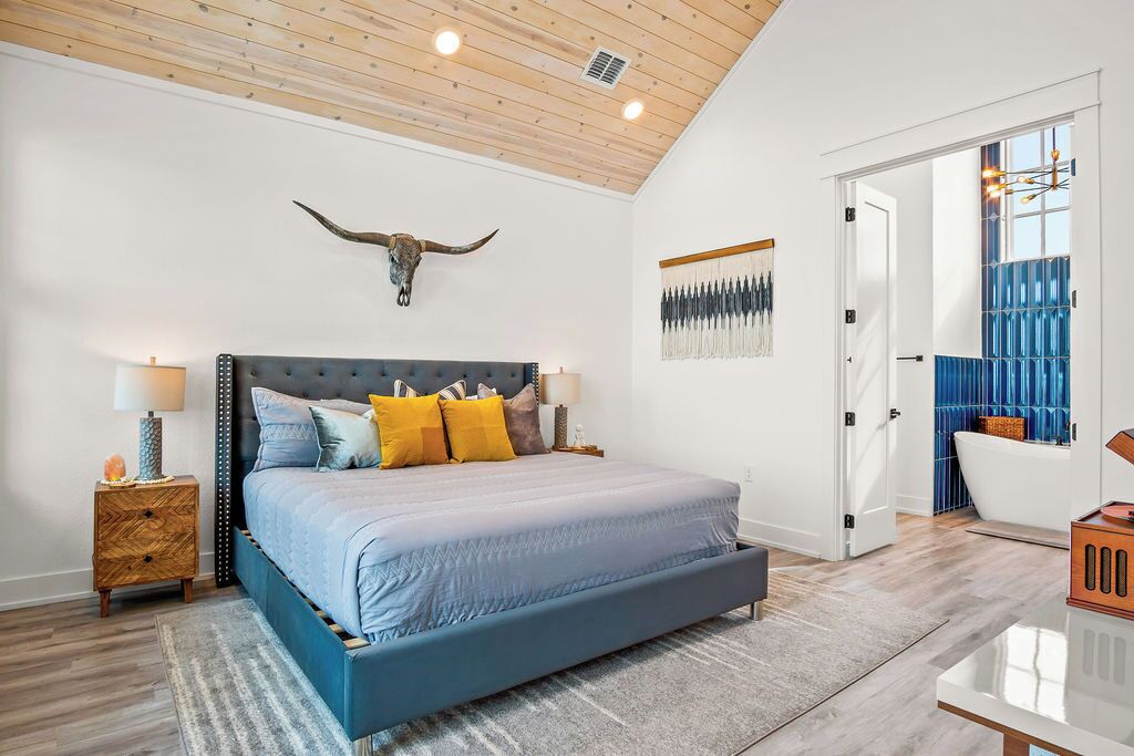 Een slaapkamer met een groot bed, een houten plafond, een decoratie van een stierenhoofd, een blauw-gele dekbedovertrek, een nachtkastje en een zichtbare badkamer door een open deur.