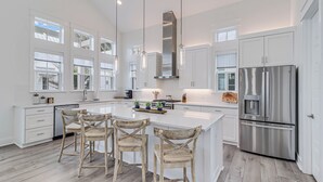 A modern kitchen with white cabinetry, a large island with bar stools, stainless steel appliances, and pendant lighting. - Beach Barn 108 BAR (Port Aransas)