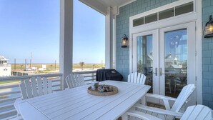 A covered outdoor area with a white table and chairs, a grill, and a view of the beach. - Still Waters 120 NEI (Port Aransas)
