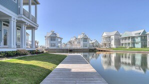 A waterfront property with a wooden dock leading to a calm body of water, surrounded by multi-story houses with balconies and a clear sky. - Still Waters 120 NEI (Port Aransas)