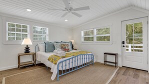 Une chambre à coucher comprenant un lit, un ventilateur de plafond, deux tables de chevet, une lampe et une porte donnant sur une vue de palmiers.