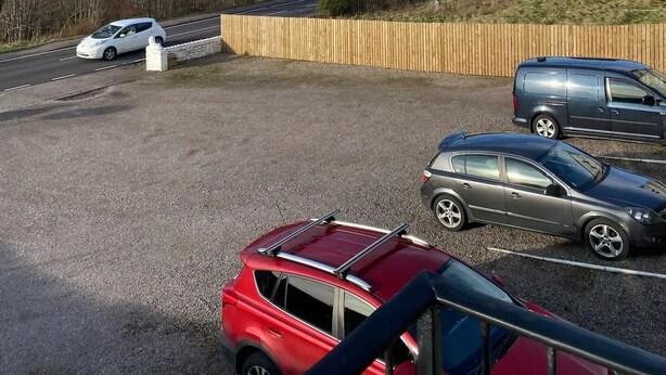 Ein Parkplatz mit mehreren Autos, einem Holzzaun und Blick auf einen See mit Bergen im Hintergrund.