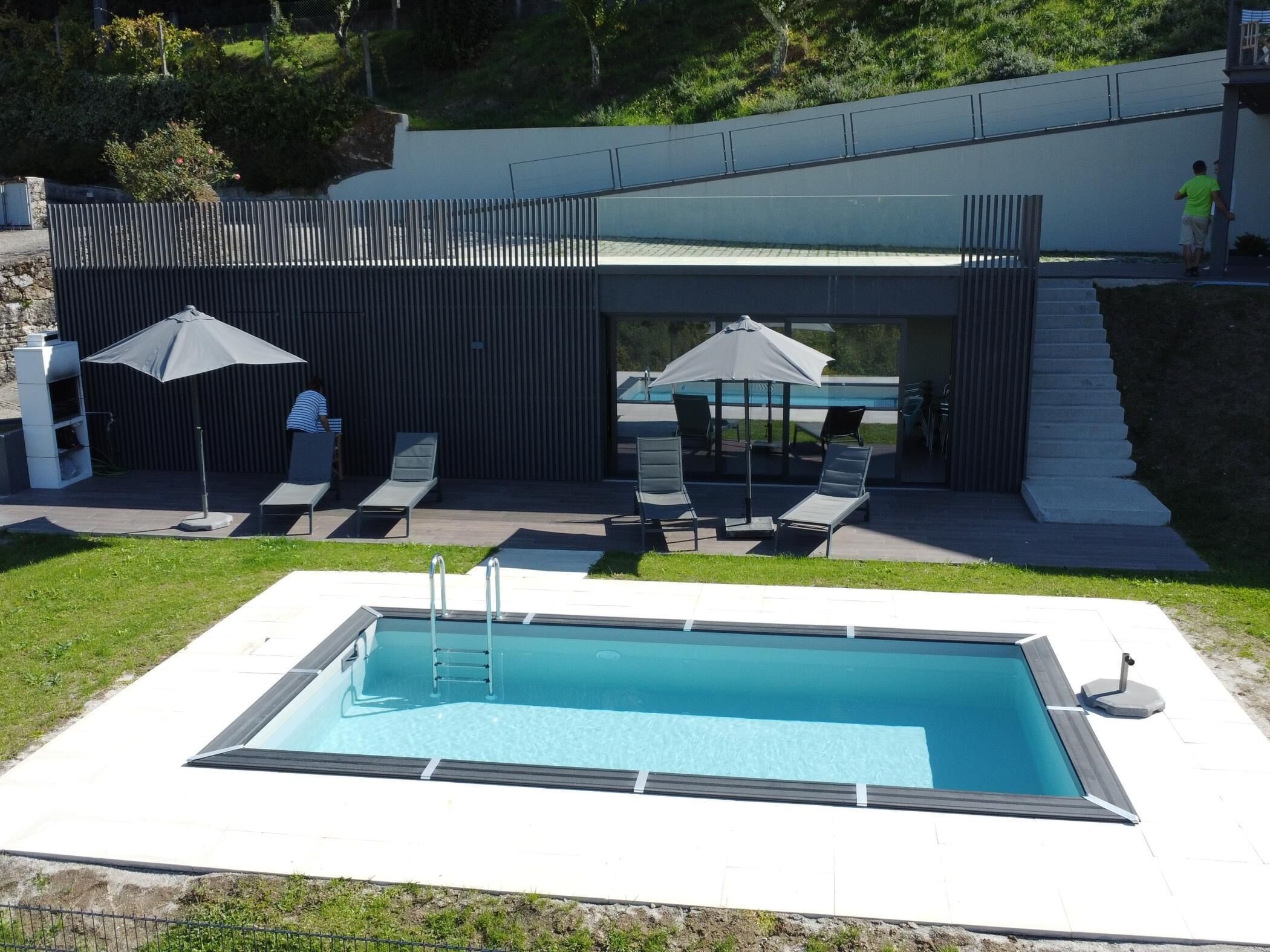 Un área de piscina moderna al aire libre con piscina rectangular, sillas de descanso y sombrillas.