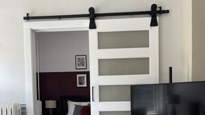 A bedroom with a bed, a TV on a tripod, and a sliding door leading to another room. - Squareview Stays - Modern Apartment On the Square (Indianola)