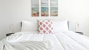 A neatly made bed with white bedding, a pink and white patterned pillow, and a white throw blanket. There are two bedside tables with lamps. Above the bed, there are three wall-mounted paintings depicting a beach scene. - Sleepover | Extraordinary 1BD/1BA - Cincinnati (Cincinnati)