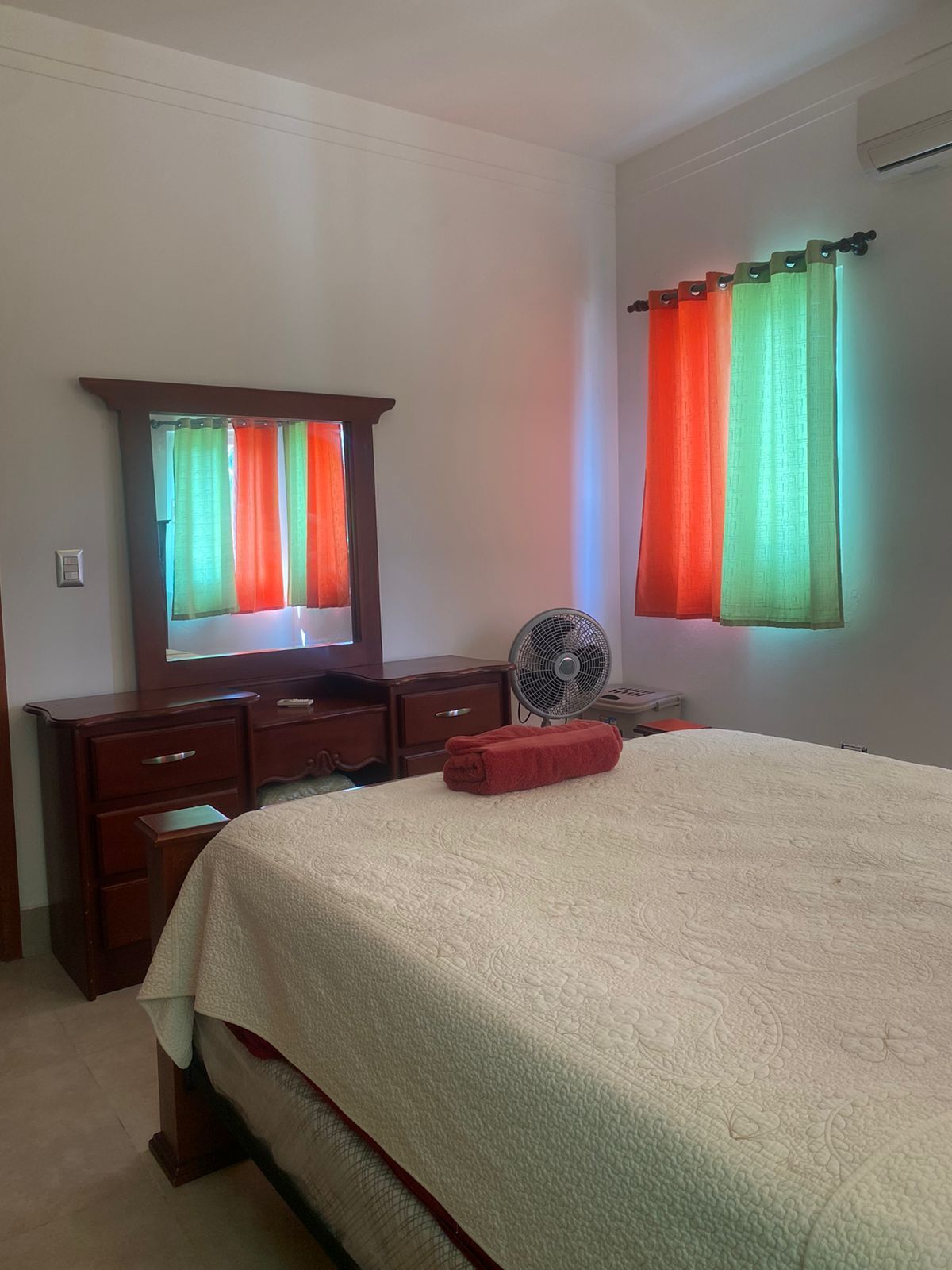 Quarto com cama, penteadeira, espelho, ventoinha e cortinas.