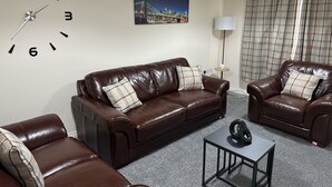 Een woonkamer met een bruine leren bank, twee fauteuils, een bijzettafeltje, een wandklok, een schilderij, een vloerlamp en ruitjesgordijnen.