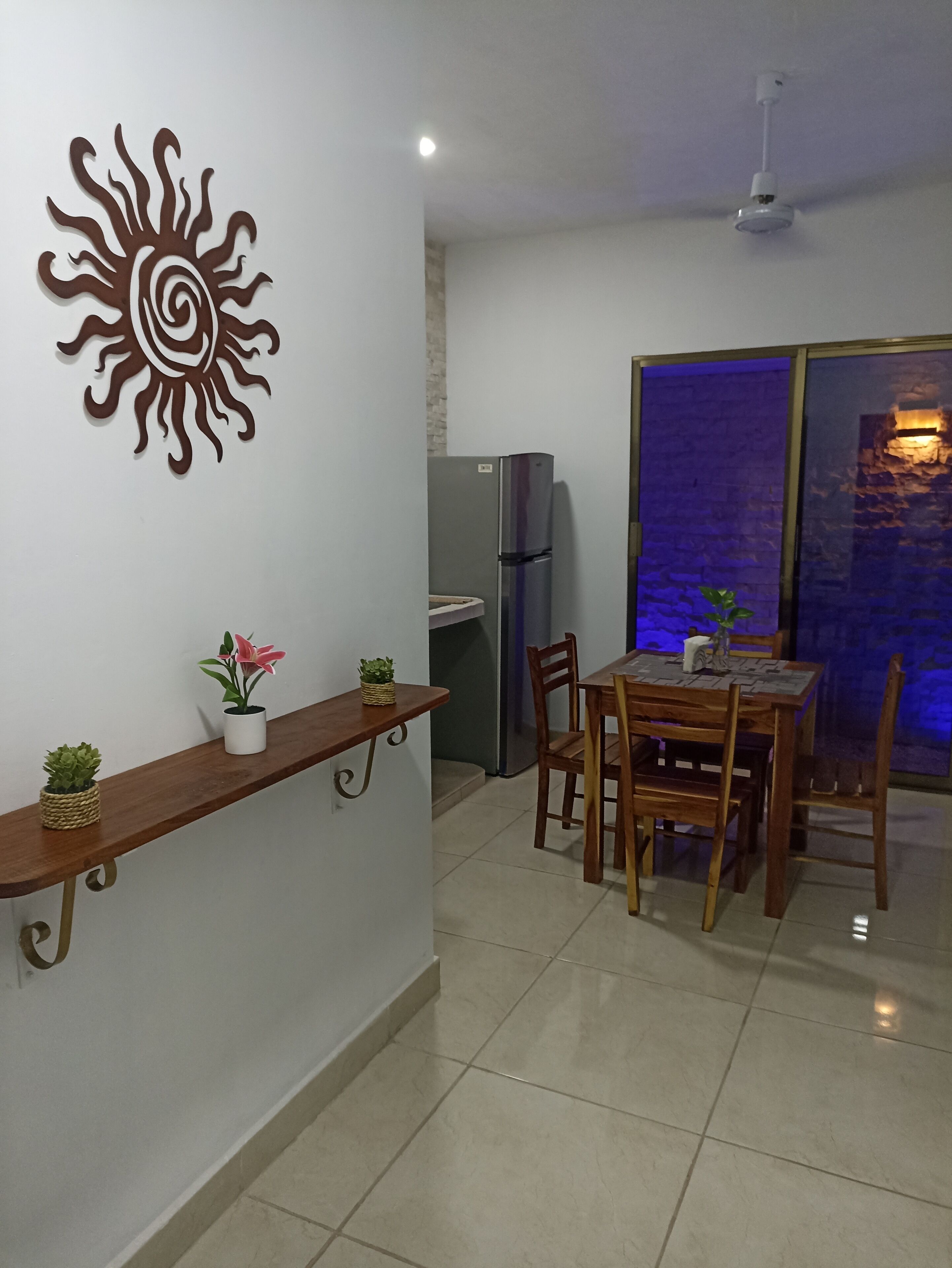 Un comedor con una mesa y sillas de madera, un refrigerador, una planta en maceta y un adorno en la pared.