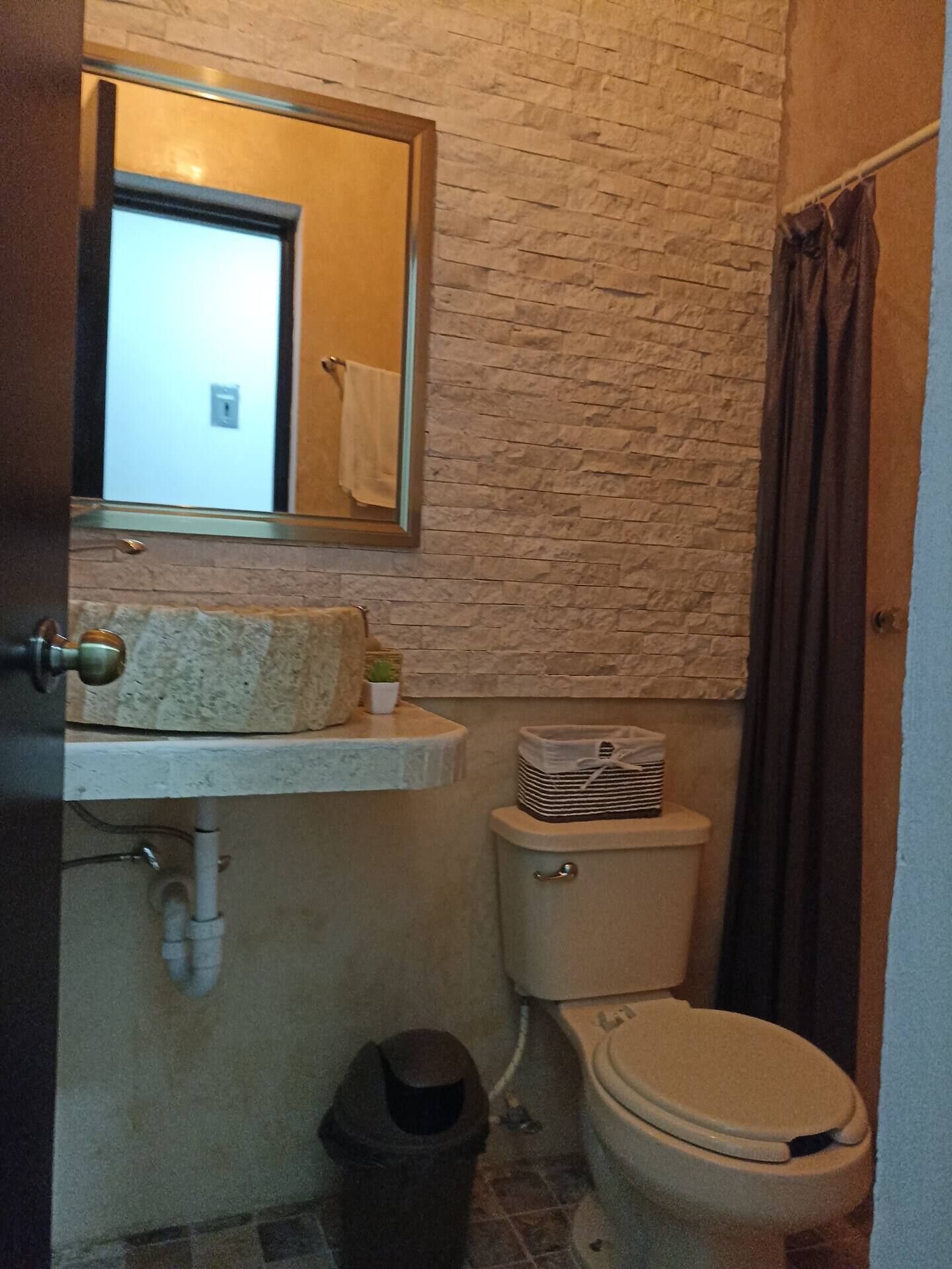 Un baño con pared de piedra, un lavabo con espejo, inodoro y cubo de basura.