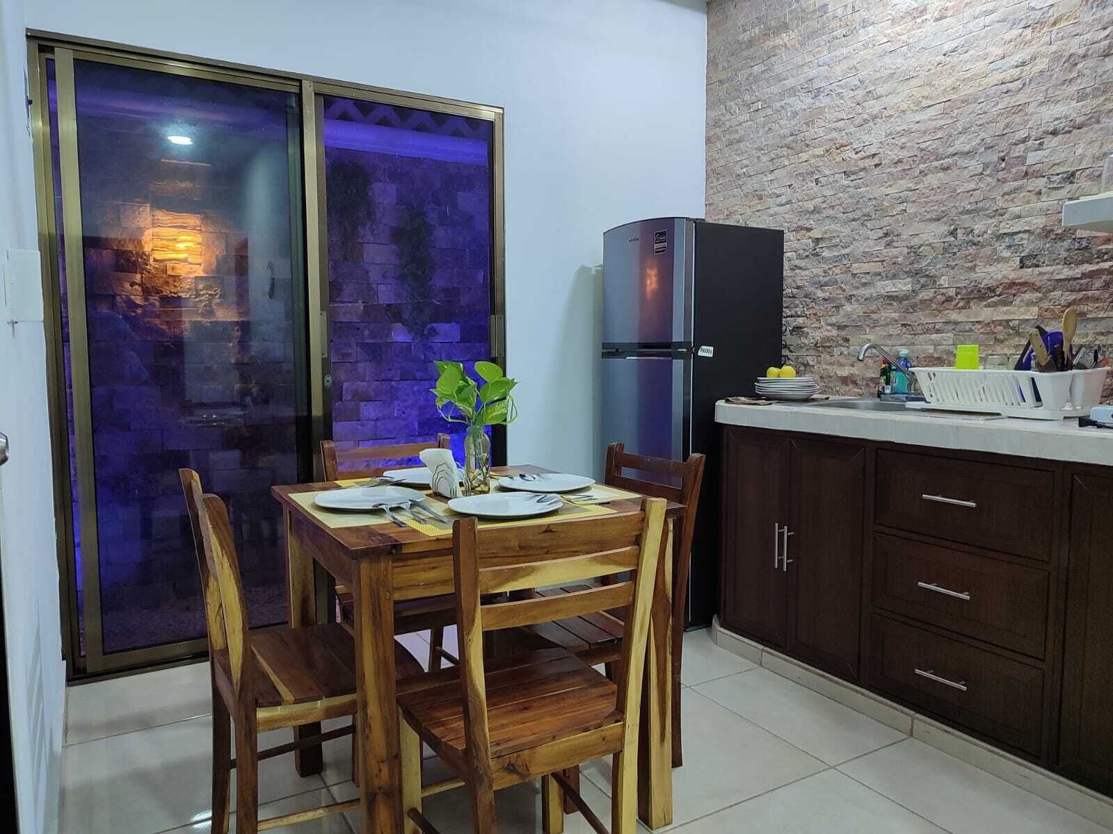 A dining area with a wooden table and chairs, a refrigerator, and a kitchen counter with cabinets.