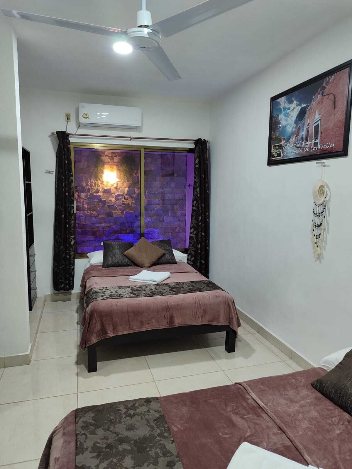 Un dormitorio con cama, ventilador de techo, aire acondicionado instalado en la pared, ventana con cortinas e imagen enmarcada en la pared.