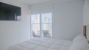 Un dormitorio con una cama grande, un televisor montado en la pared y vistas de edificios altos a través de la ventana.