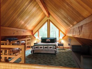 Une chambre mansardée chaleureuse, avec un plafond en pente, un lit recouvert d’une couette à motifs d’ours, une fenêtre donnant sur les arbres et un ventilateur fixé au mur.