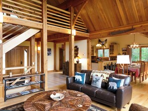A spacious living area with a wooden ceiling, a leather sofa with animal-themed cushions, a round coffee table, and a staircase leading to an upper level.