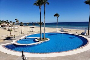 Una piscina de borde curvo, rodeada de palmeras y una playa al fondo.