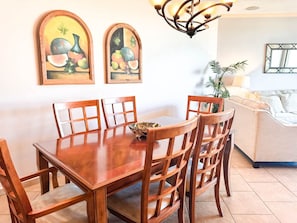 A dining area with a wooden table and chairs, two framed paintings, a chandelier, and a sofa in the background.