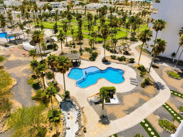An aerial view of a resort with a central swimming pool, palm trees, and a well-maintained landscape.