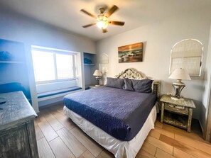 A bedroom with a bed, a ceiling fan, a window with blinds, a bedside table with a lamp, and a wall-mounted shelf.