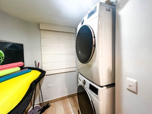 Uma lavanderia compacta com máquina de lavar e secadora, uma janela com persianas e uma pequena mesa com produtos de limpeza.