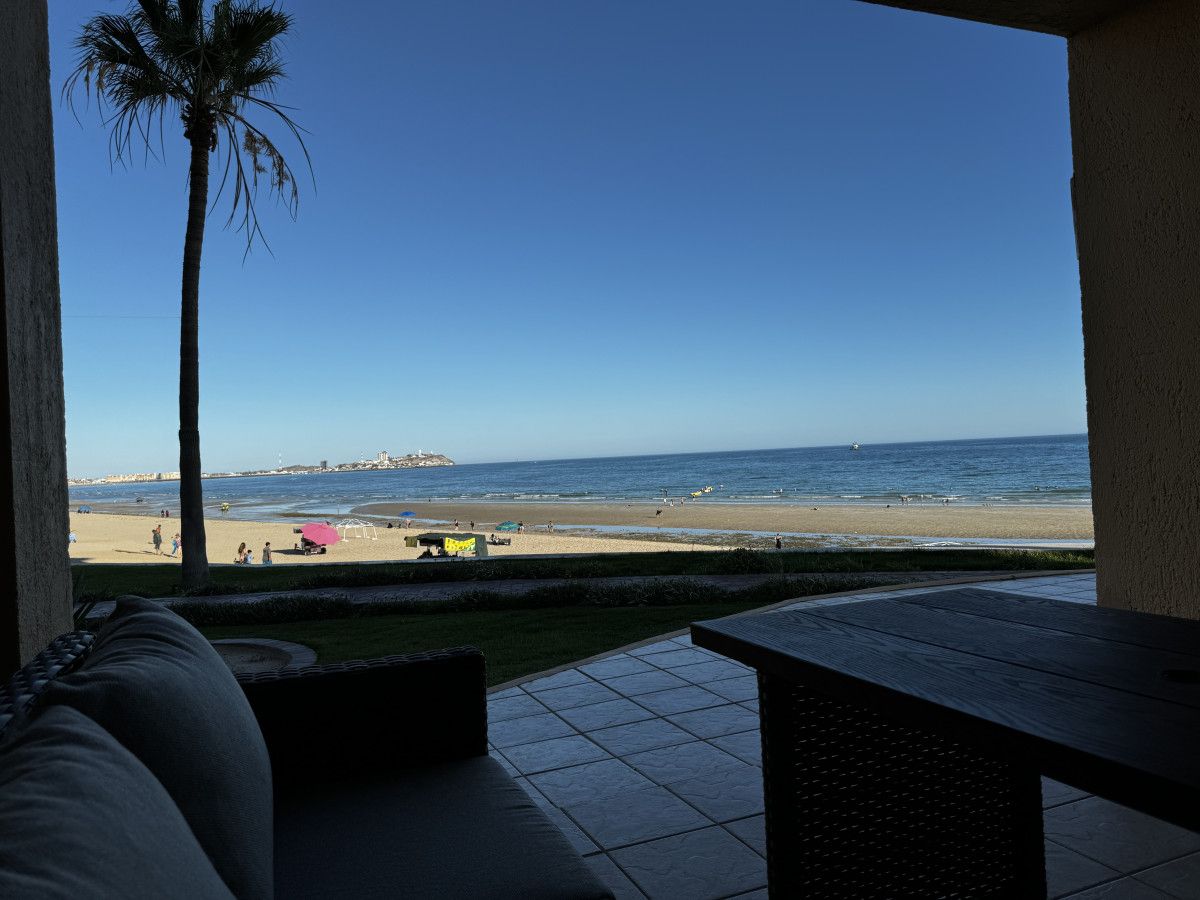 A patio with a view of a beach, a palm tree, and a distant shoreline.