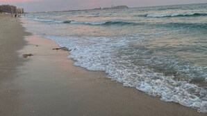 Une plage où les vagues viennent doucement lécher le rivage, un ciel dégagé et des bâtiments qui se dessinent à l’horizon.