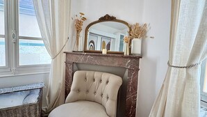 A room with a marble fireplace, a cushioned armchair, a wicker basket, and a mirror above the fireplace.