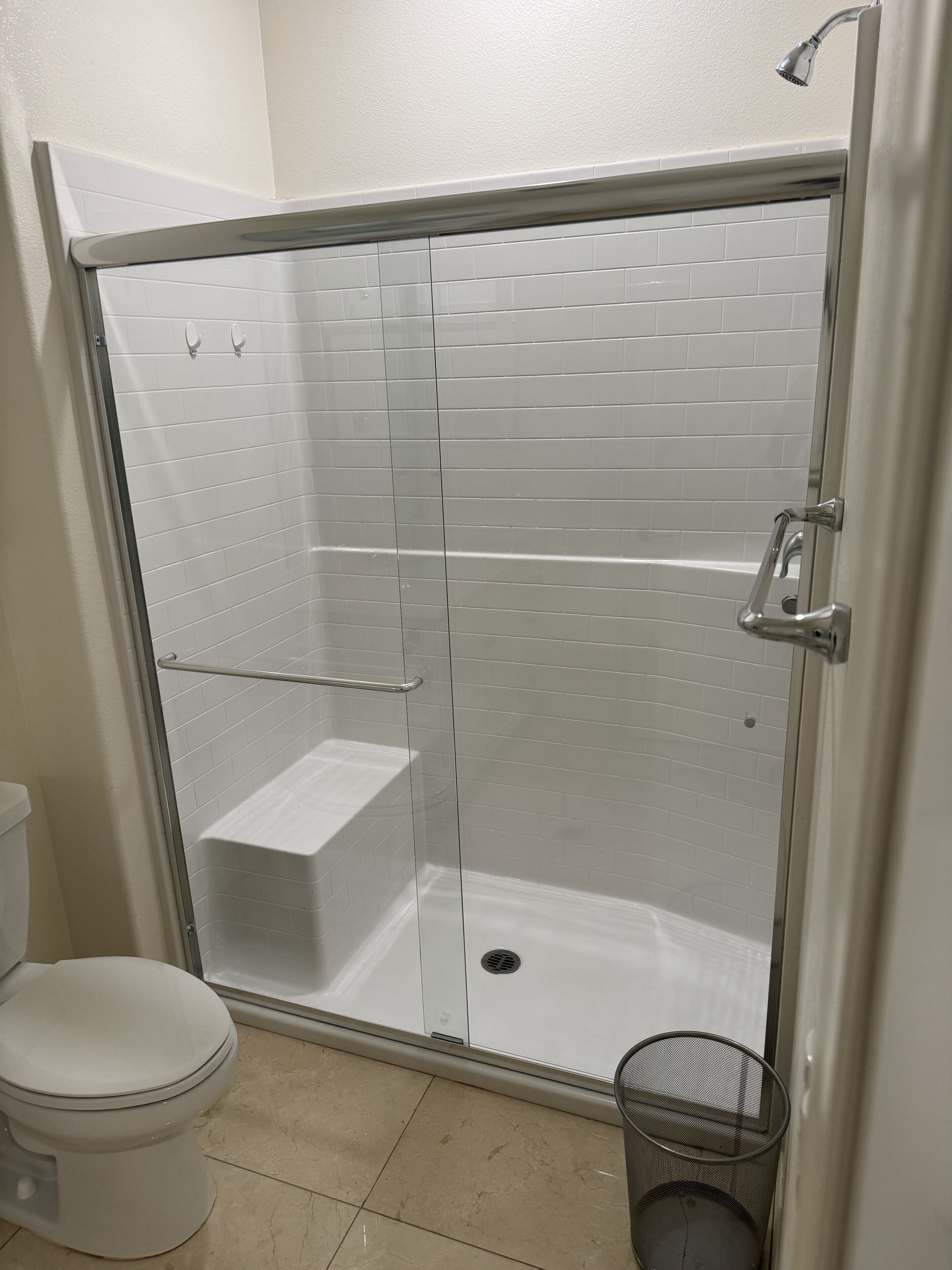 A glass-enclosed shower stall with white tiled walls and a white toilet to the left.