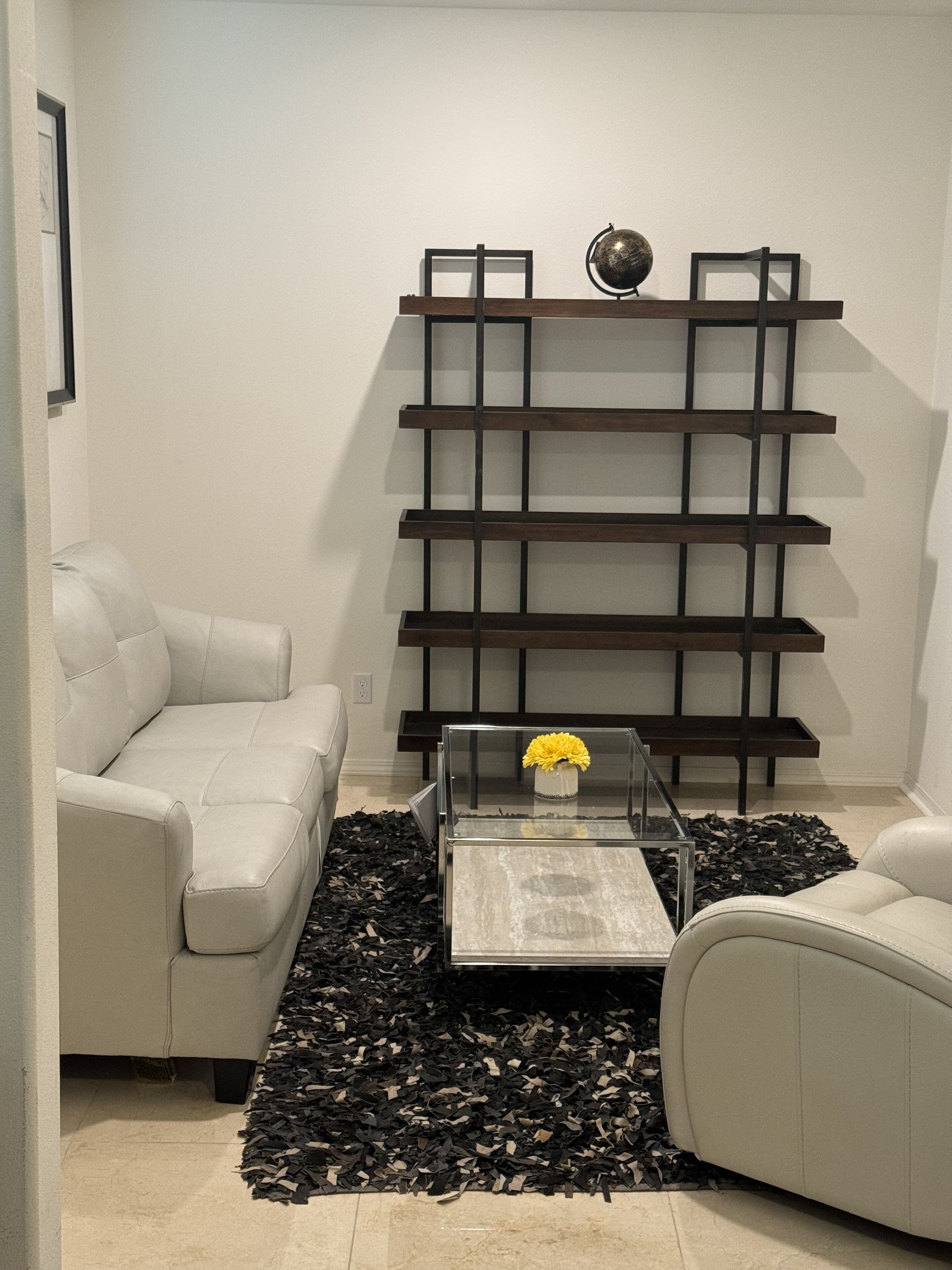 A modern living room with a sectional sofa, a glass coffee table with a yellow flower, and a dark wooden shelving unit against the wall.