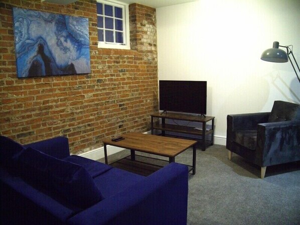 Sala de estar con pared de ladrillo, televisor de pantalla plana, sofá azul oscuro, una mesa de centro y una lámpara de pie.