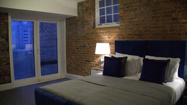 A bedroom with a large bed, a nightstand, a lamp, a window with blinds, and a brick wall. - Old Dutch Embassy by Guestz (Kingston-upon-Hull)
