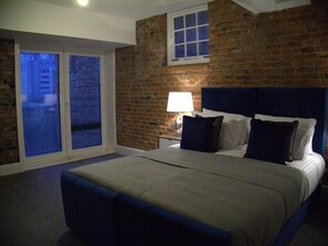 Un dormitorio con una cama grande, una mesita de noche, una lámpara, una ventana con persianas y una pared de ladrillo.