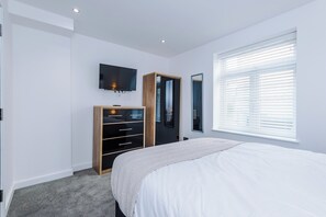 A bedroom with a bed, a TV mounted on the wall, a wardrobe, and a window with blinds.