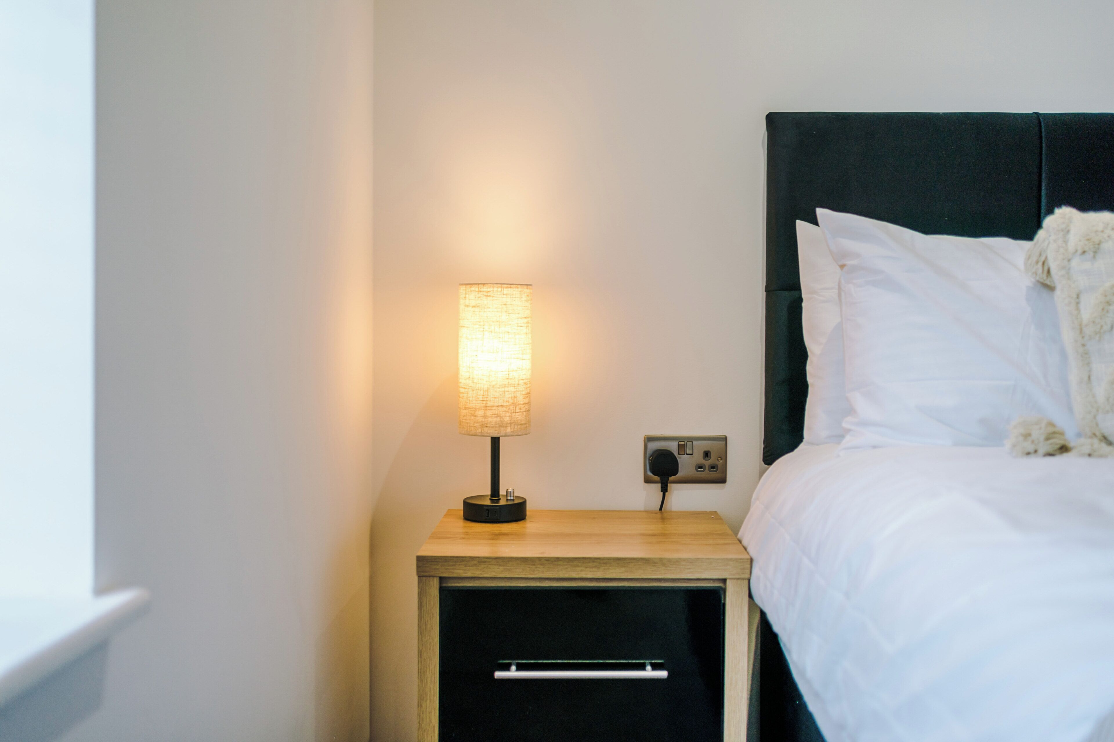 A neatly made bed with white linens, a bedside lamp, and a small wooden nightstand with a drawer.