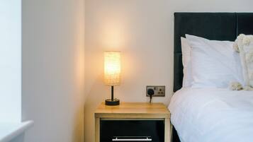 A neatly made bed with white linens, a bedside lamp, and a small wooden nightstand with a drawer.