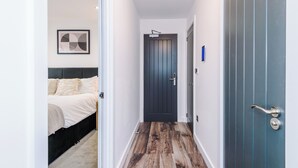 Un pasillo estrecho con una cama a la izquierda, una puerta oscura al final y una puerta azul a la derecha.