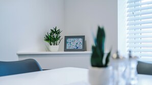 Una habitación moderna con una mesa blanca, una planta en maceta, una obra de arte enmarcada y una ventana con persianas.