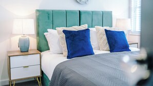Un letto ben rifatto con una testiera verde, biancheria da letto bianca e cuscini blu e beige. È presente un comodino con una lampada e un elemento decorativo sopra il letto.
