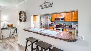 Cocina con armarios de madera, refrigerador de acero inoxidable y microondas. Hay dos taburetes de bar y una encimera con papeles.