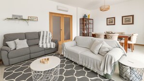 Un salon comprenant un canapé gris, un canapé blanc, une table basse ronde et un coin repas avec une table et des chaises en bois.
