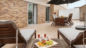 Une terrasse avec une table en bois dressée pour deux personnes, une chaise en bois, un grand parasol et un plateau de fruits et de boissons.