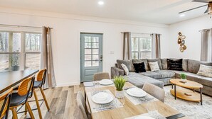A modern living room with a dining area, a grey sofa set, and a wooden dining table set for four. - Winnsboro Barndominium w/ Fire Pit & Large Deck! (Winnsboro)