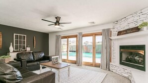 A living room with a fireplace, a ceiling fan, and a view of a pool. - Lakeview w/Pool, Hot Tub, Firepit, Gameroom, Golf! (Penn Yan)